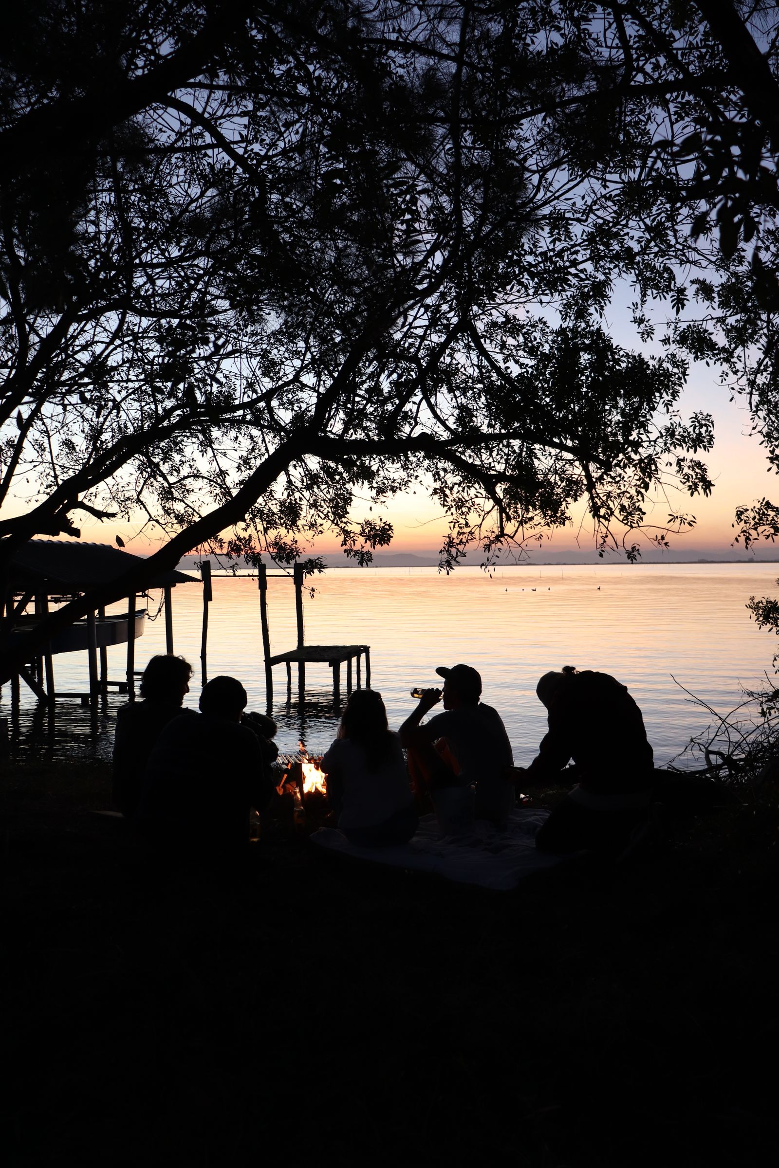 Friends gathered around a small fire beside a calm lake at dusk
